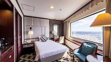 Ein Hotelzimmer mit einem großen Bett, einem Schreibtisch, einem Stuhl, Blick auf die Stadt und einem mit Vorhängen versehenen Fenster.