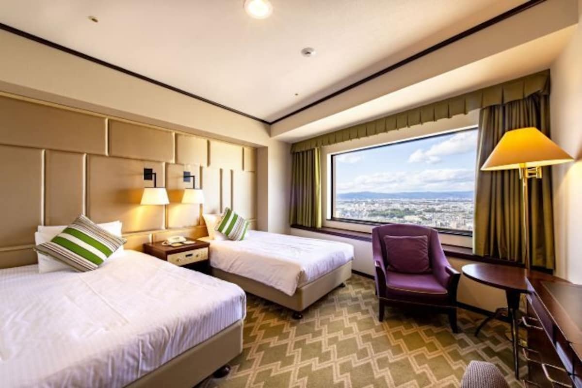 A hotel room with two beds, a chair, a small table, a lamp, and a large window offering a city view.