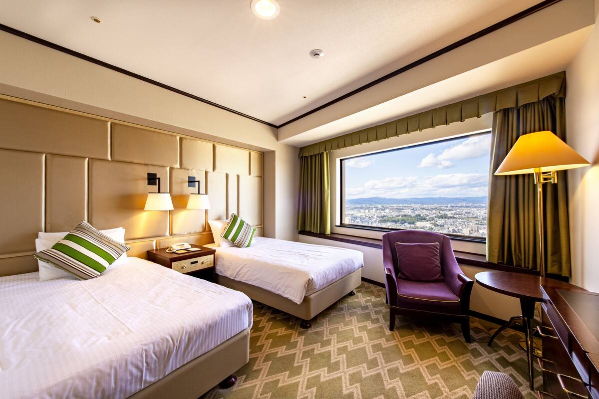 A hotel room with two beds, a chair, a desk, and a view of the city.
