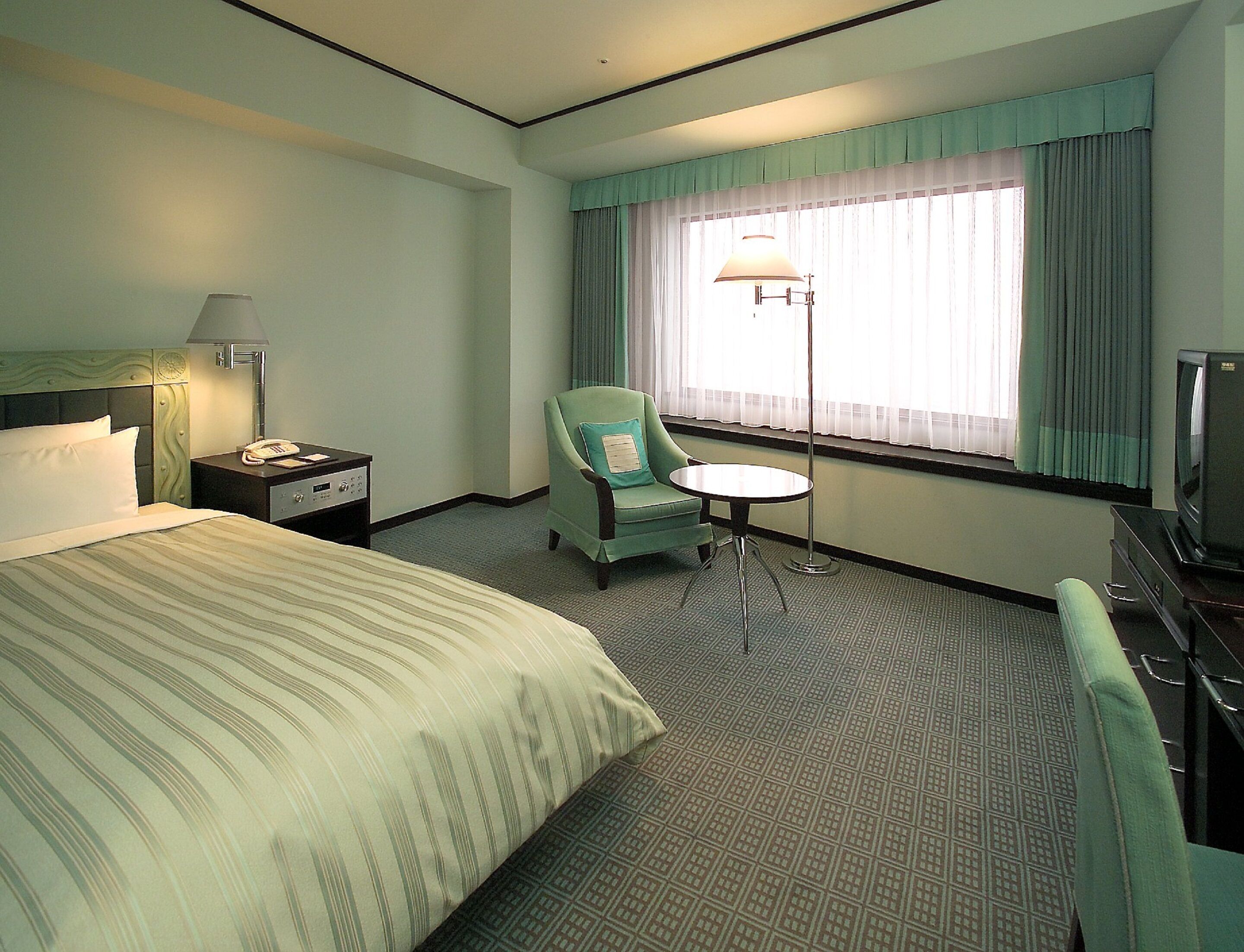 A hotel room with a bed, a chair, a small table, a lamp, a television, and a window with curtains.