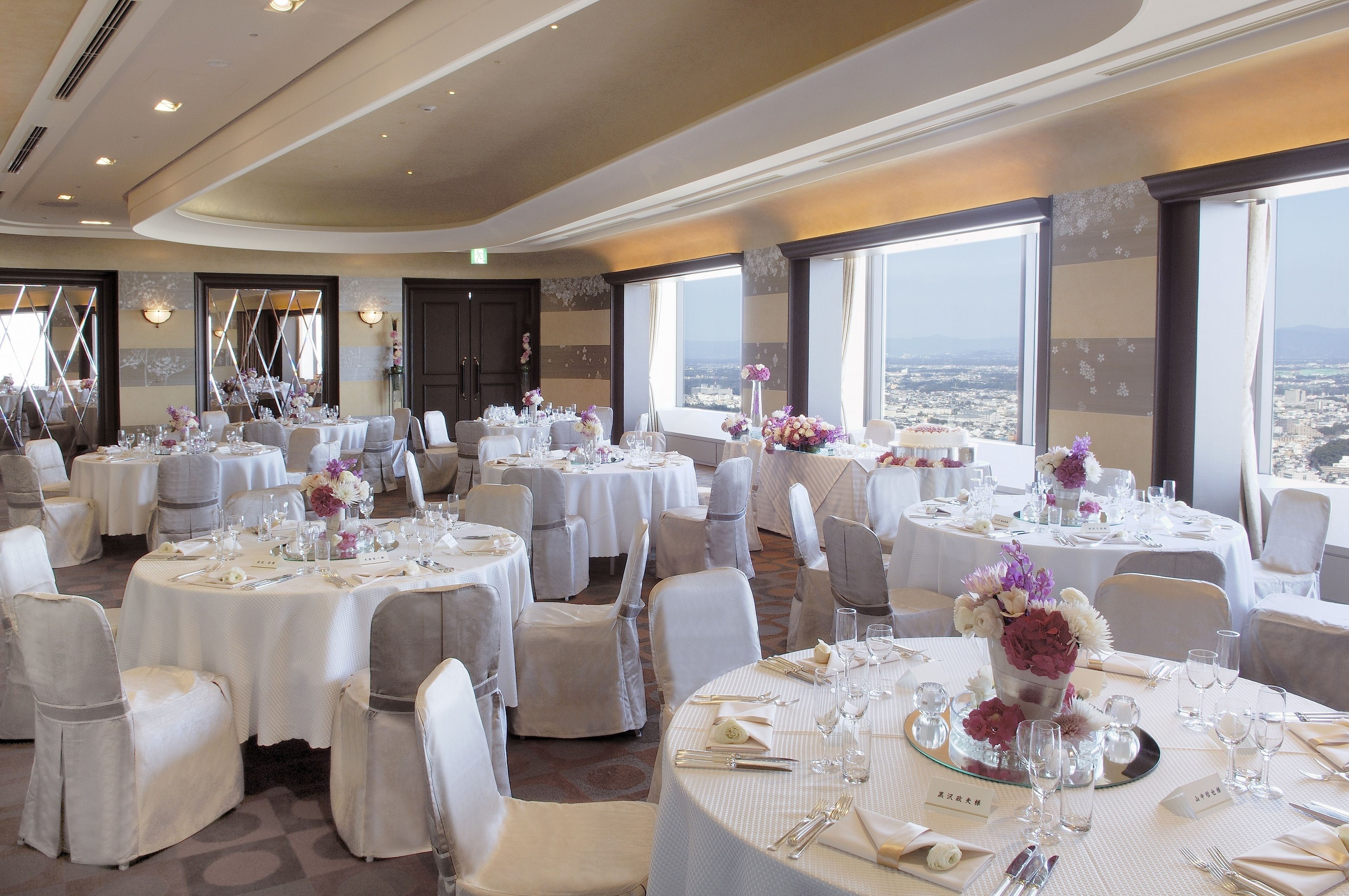 A banquet hall with round tables set for dining, large windows offering a city view, and a high ceiling with recessed lighting.