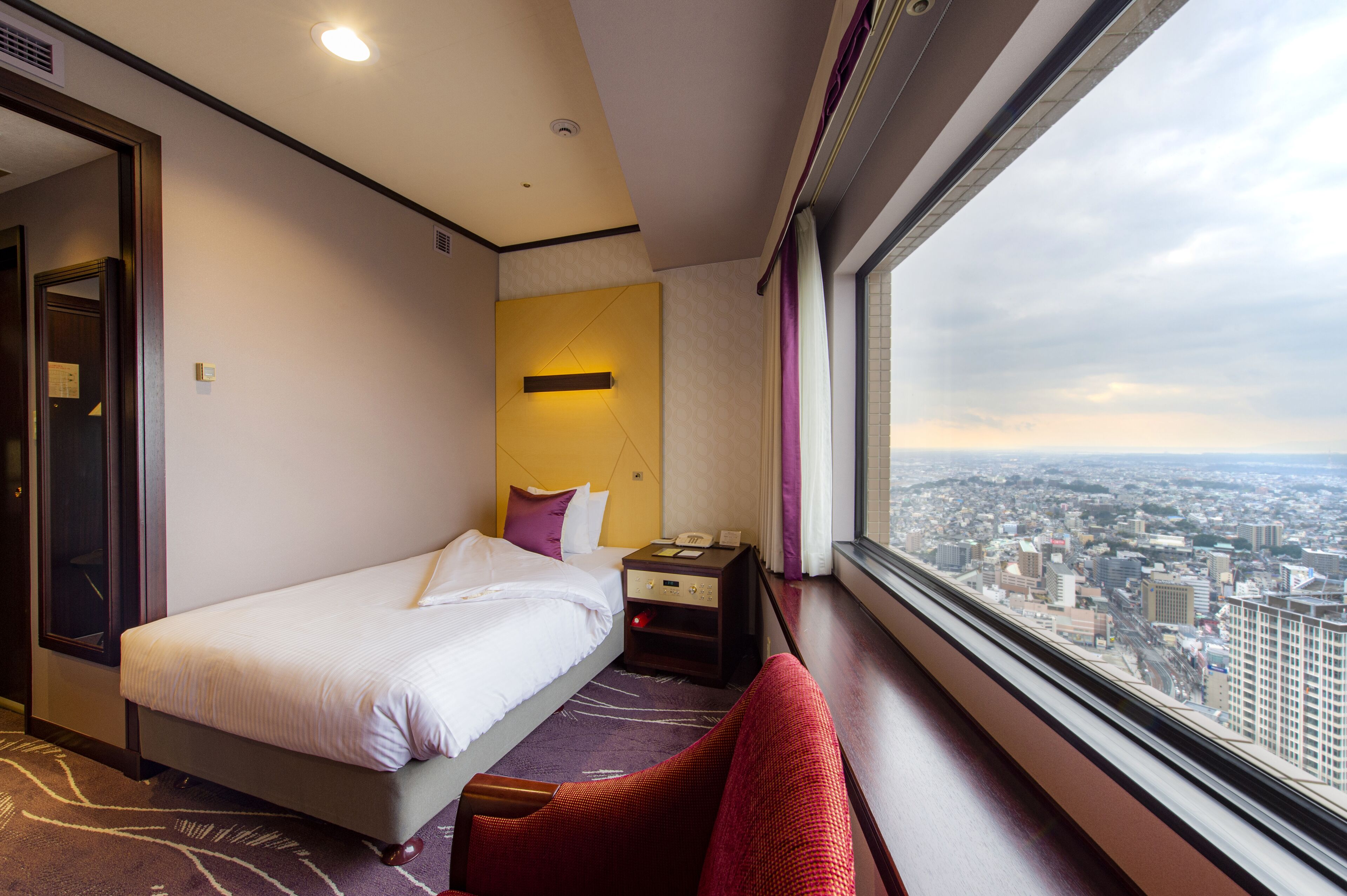A hotel room with a bed, a nightstand, a window with a city view, and a red chair.