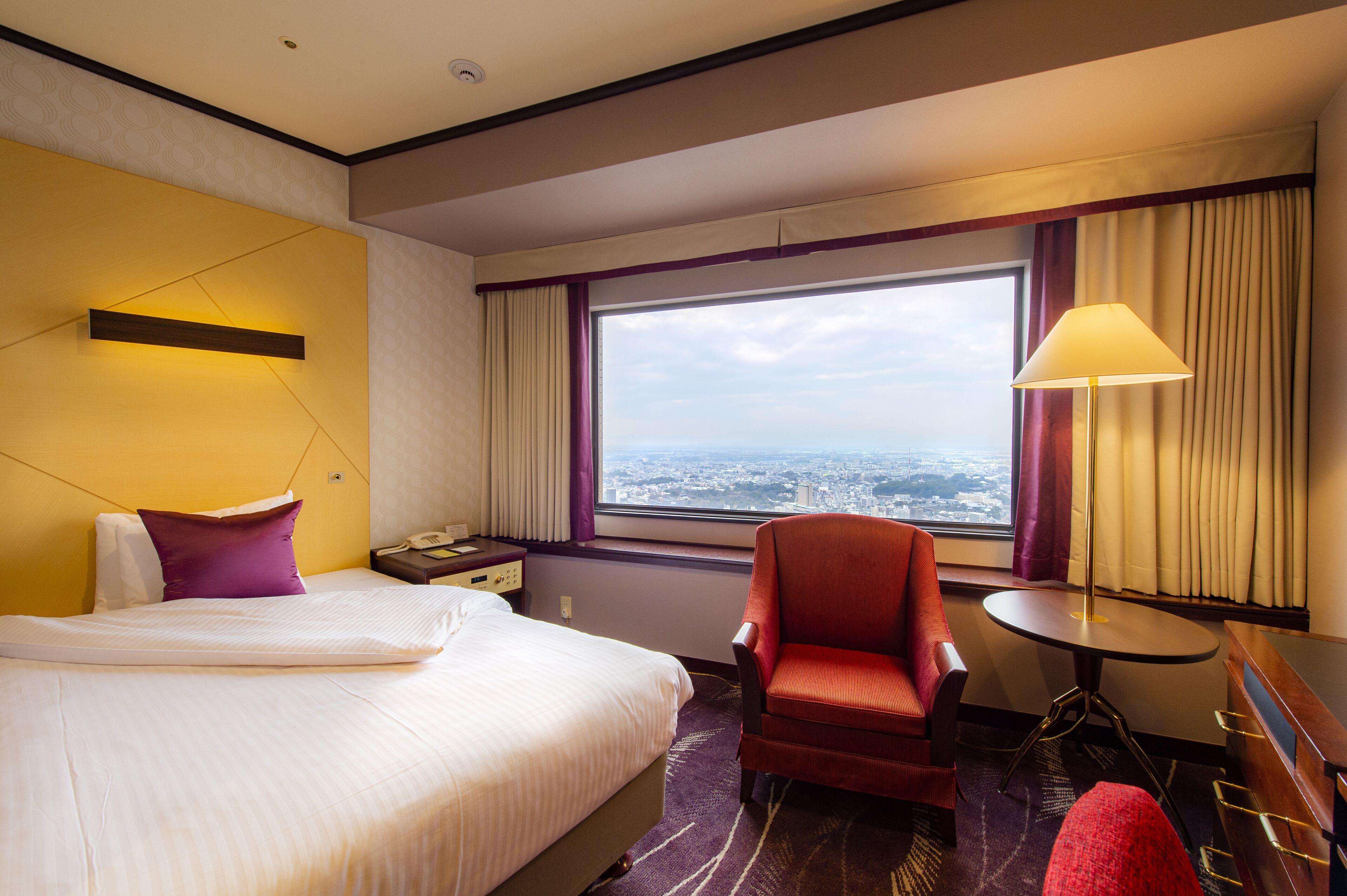A hotel room with a bed, a red armchair, a small round table, and a view of the city through the window.