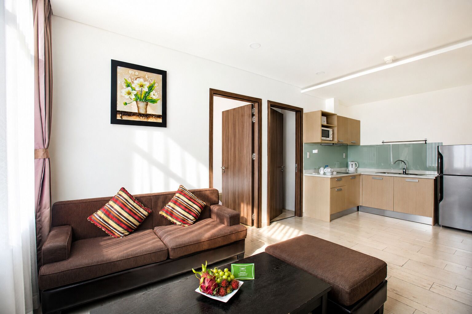 A modern apartment with a sofa, a coffee table with fruit, a kitchen with a microwave, and a door leading to another room.