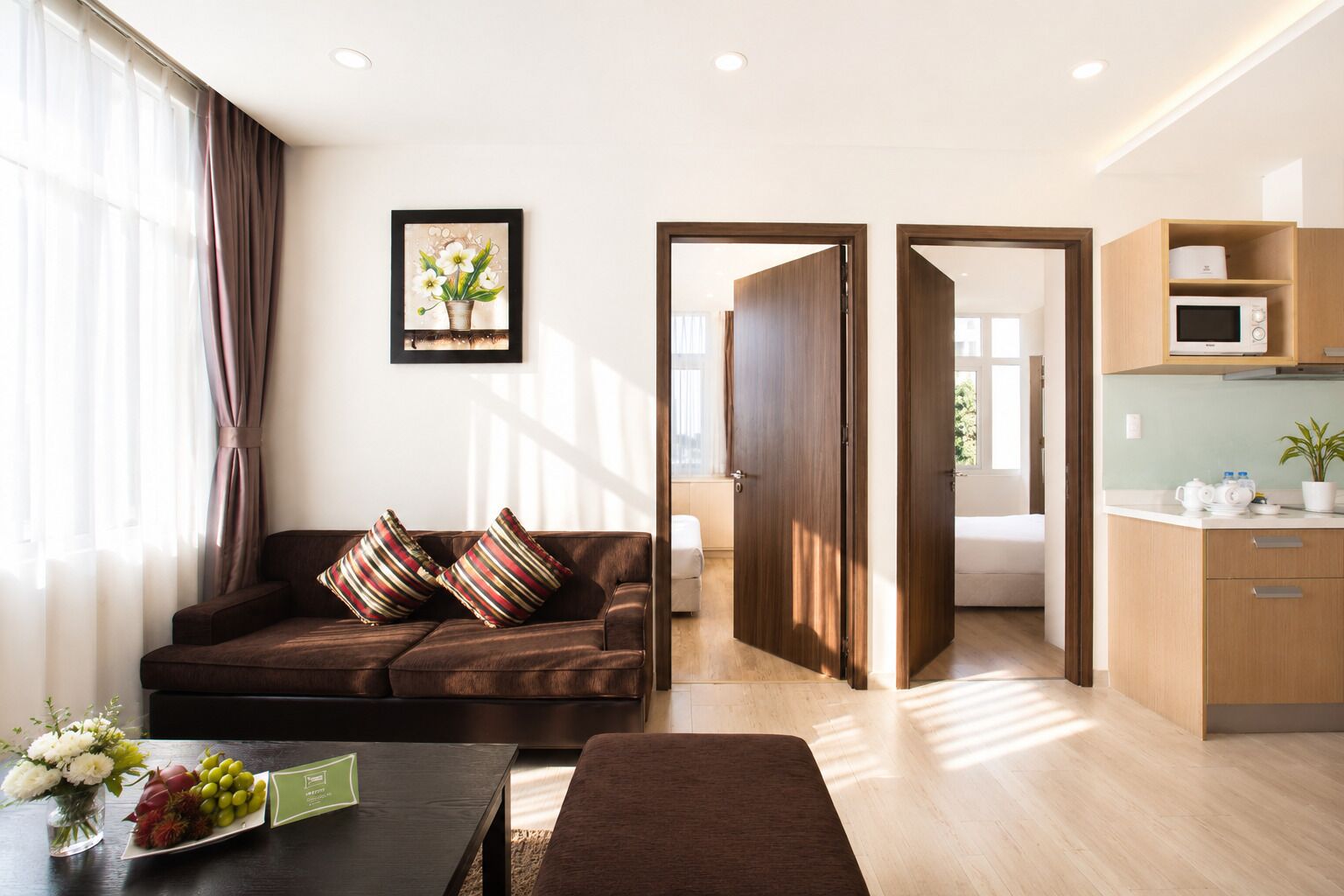 A modern living room with a sofa, a dining table with a vase of flowers, a kitchen area with a microwave, and a view through an open door.