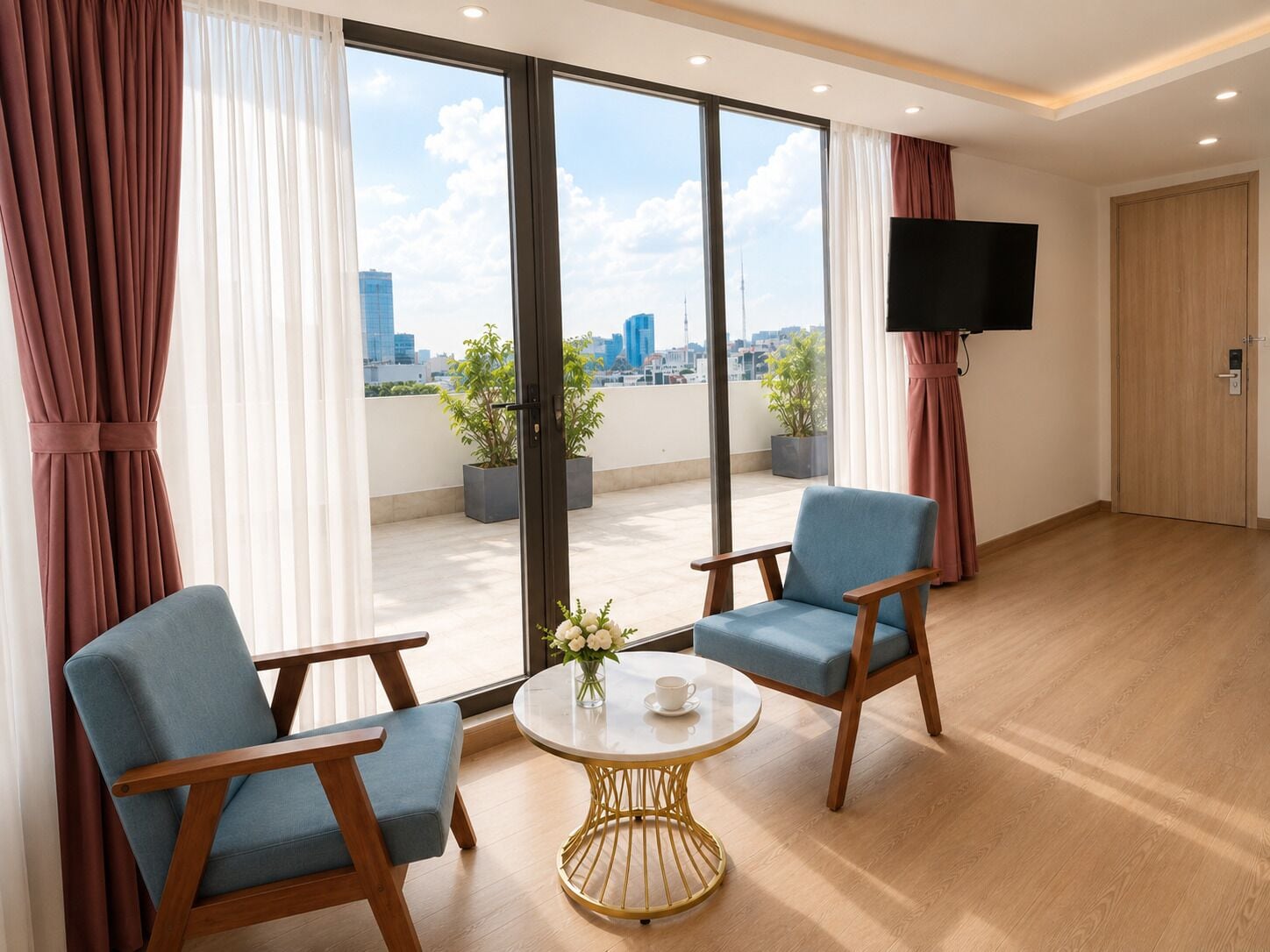 A hotel room with two blue armchairs, a round table with a gold base, a flat-screen TV mounted on the wall, and a city view through large windows.