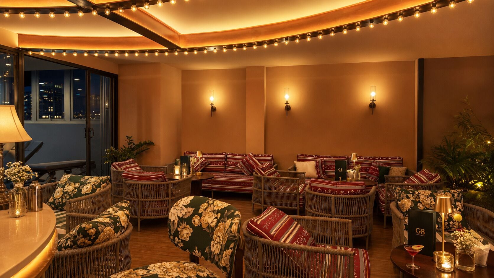 A cozy lounge area with wicker furniture, wall sconces, and a view of the city at night.