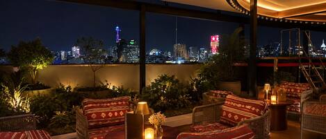 A rooftop lounge with wicker furniture, a view of a city skyline at night, and a large window.