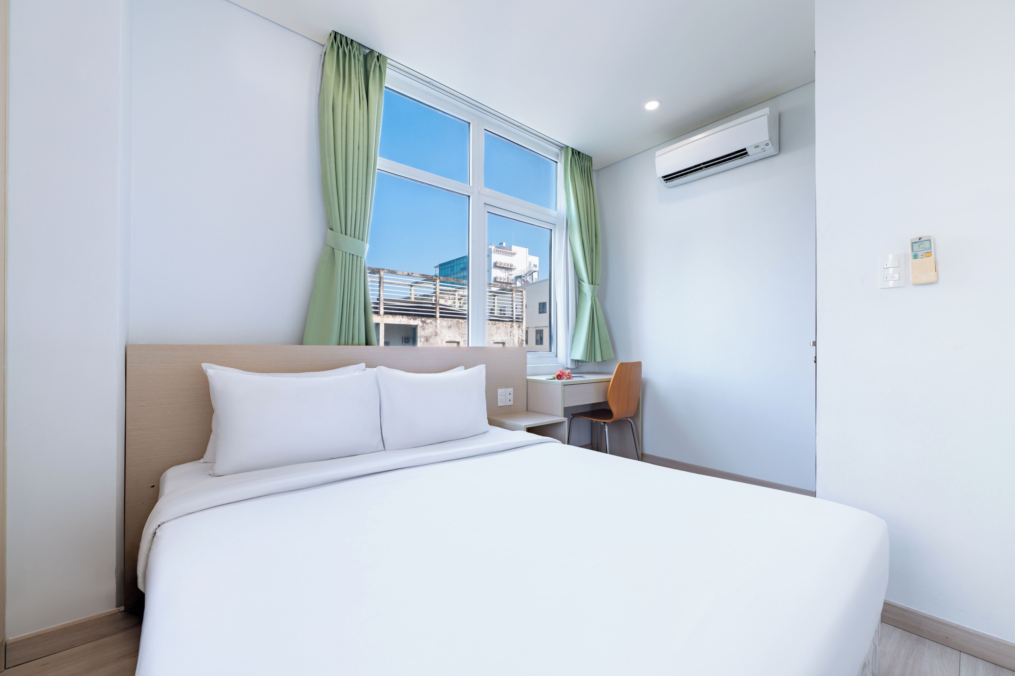 A hotel room with a bed, a desk, a chair, a window with curtains, an air conditioning unit, and a wall-mounted thermostat.