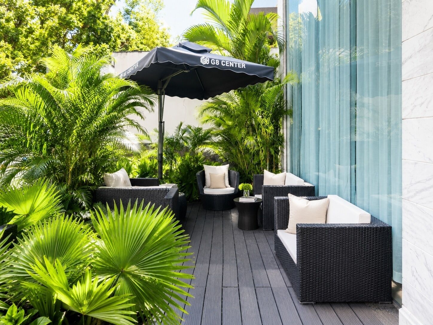 A patio area with wicker furniture, a black umbrella, and lush green plants.