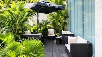 A patio area with wicker furniture, a black umbrella, and lush green plants.