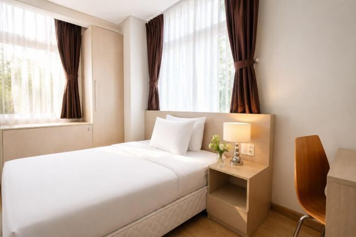 Une chambre d’hôtel avec un lit, une table de chevet avec une lampe, une chaise et une fenêtre avec des rideaux.