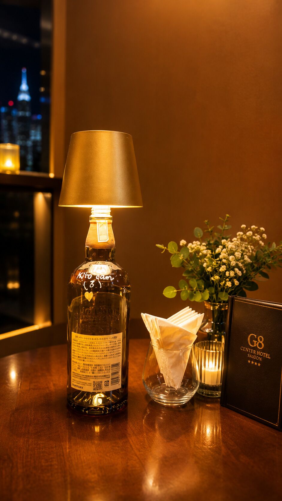 A bottle of liquor, a lamp, a vase with flowers, a napkin holder, and a sign on a wooden table.