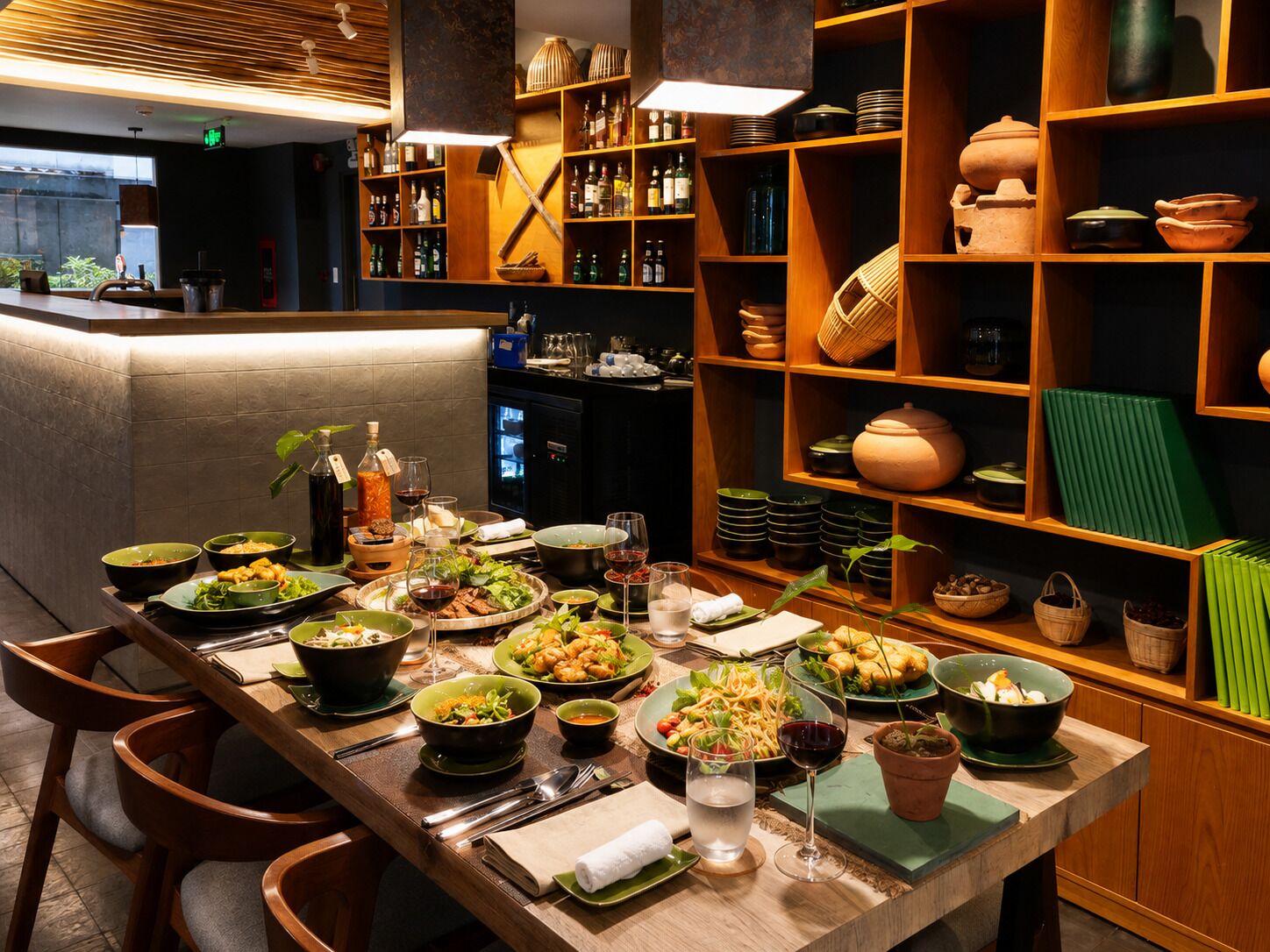Interior de restaurante con una larga mesa de comedor dispuesta para varios comensales, rodeada de estantes con utensilios de cocina y botellas.