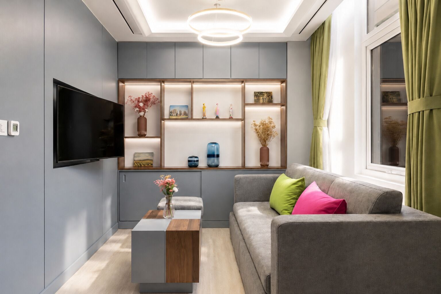 A modern living room with a flat-screen TV, a wooden coffee table, a grey sofa with colorful cushions, and a built-in shelving unit with decorative items.