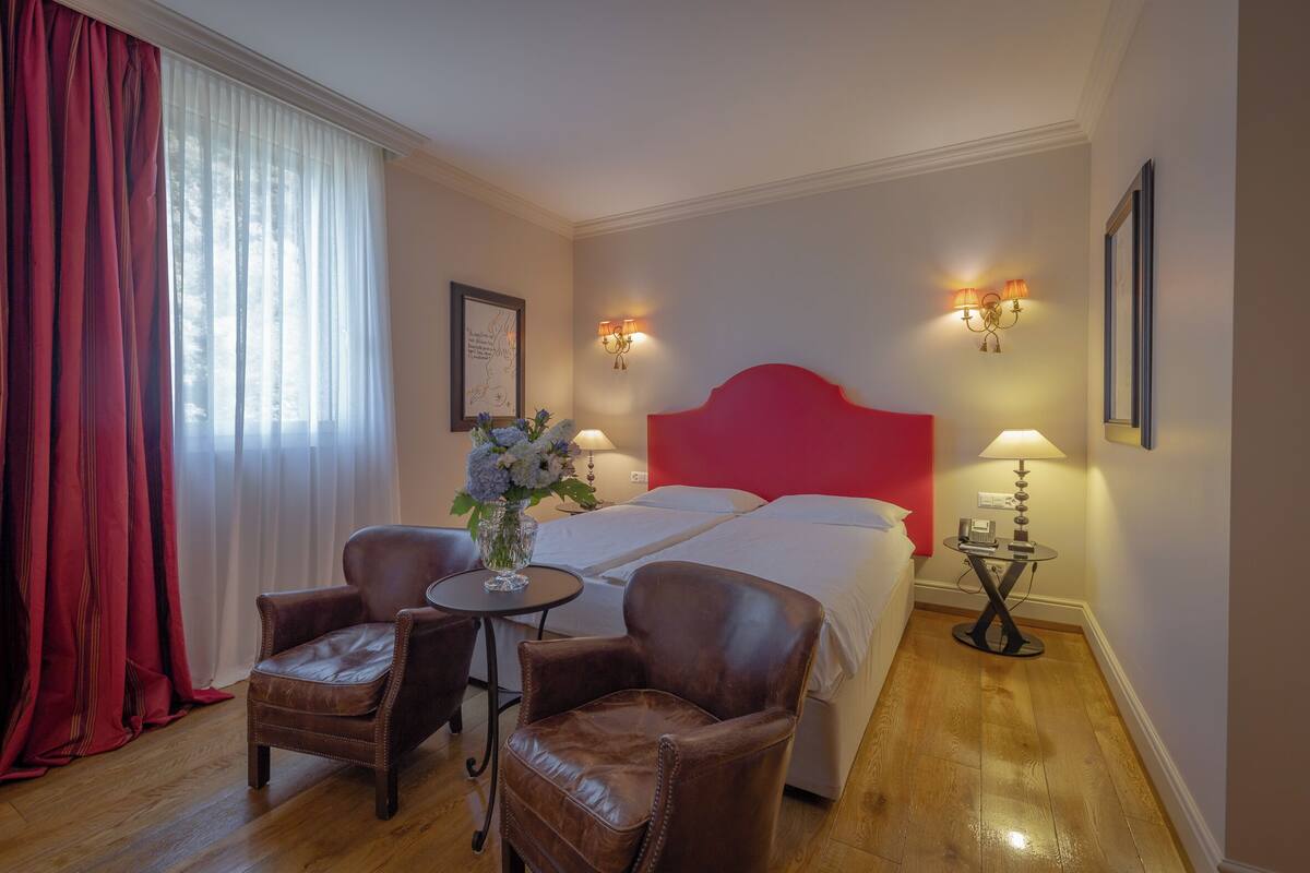 A hotel room with a bed, two chairs, a small table with a vase of flowers, and two wall-mounted lamps.