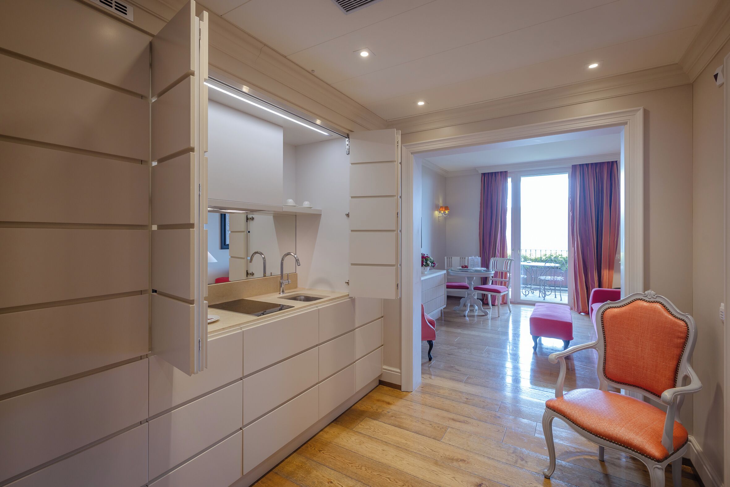A modern kitchen with integrated appliances, a dining area with a table and chairs, and a balcony with curtains.