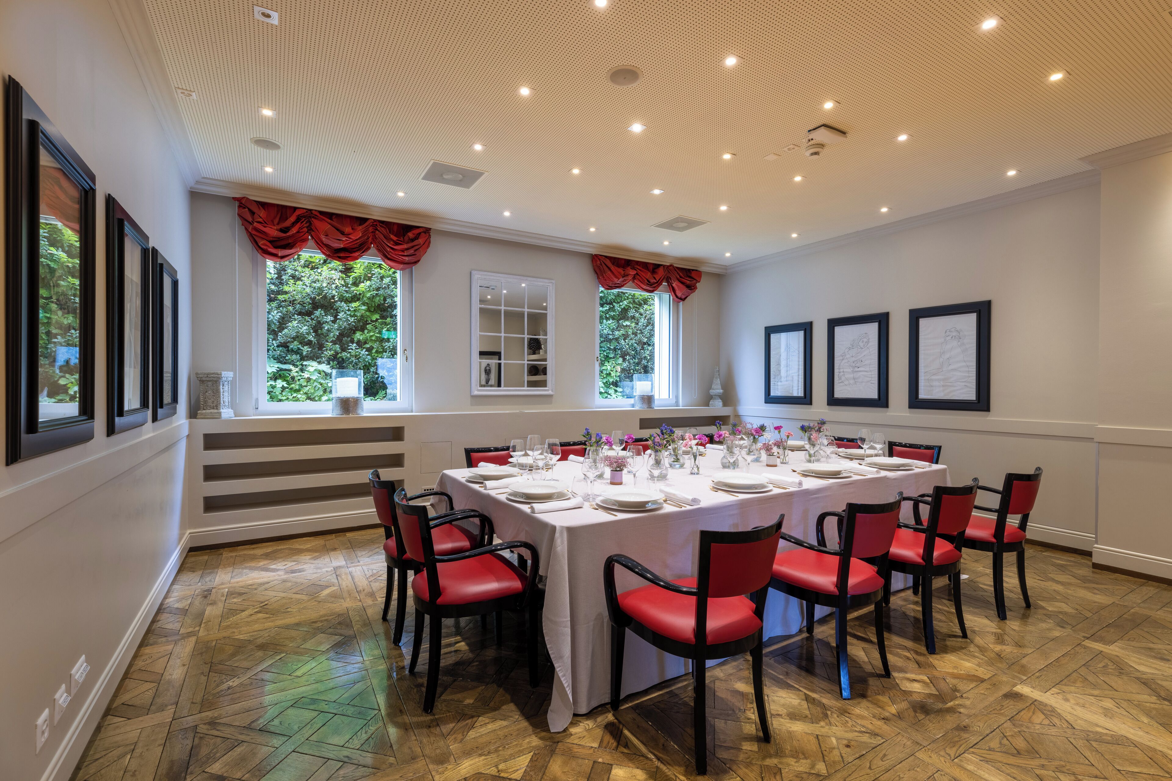 Ein Esszimmer mit einem langen Tisch, gedeckt für sechs Personen, umgeben von roten Stühlen, großen Fenstern mit roten Vorhängen und gerahmten Bildern an den Wänden.