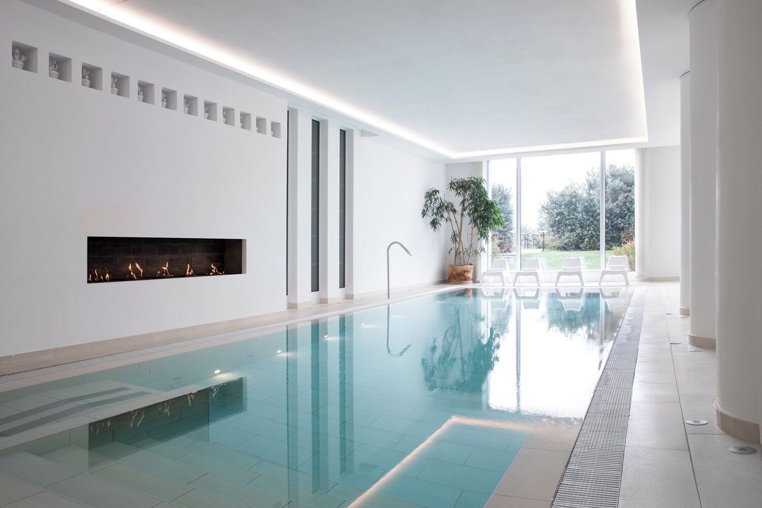 Une piscine intérieure moderne dotée d’une cheminée intégrée, de grandes fenêtres et offrant une vue sur l’extérieur.