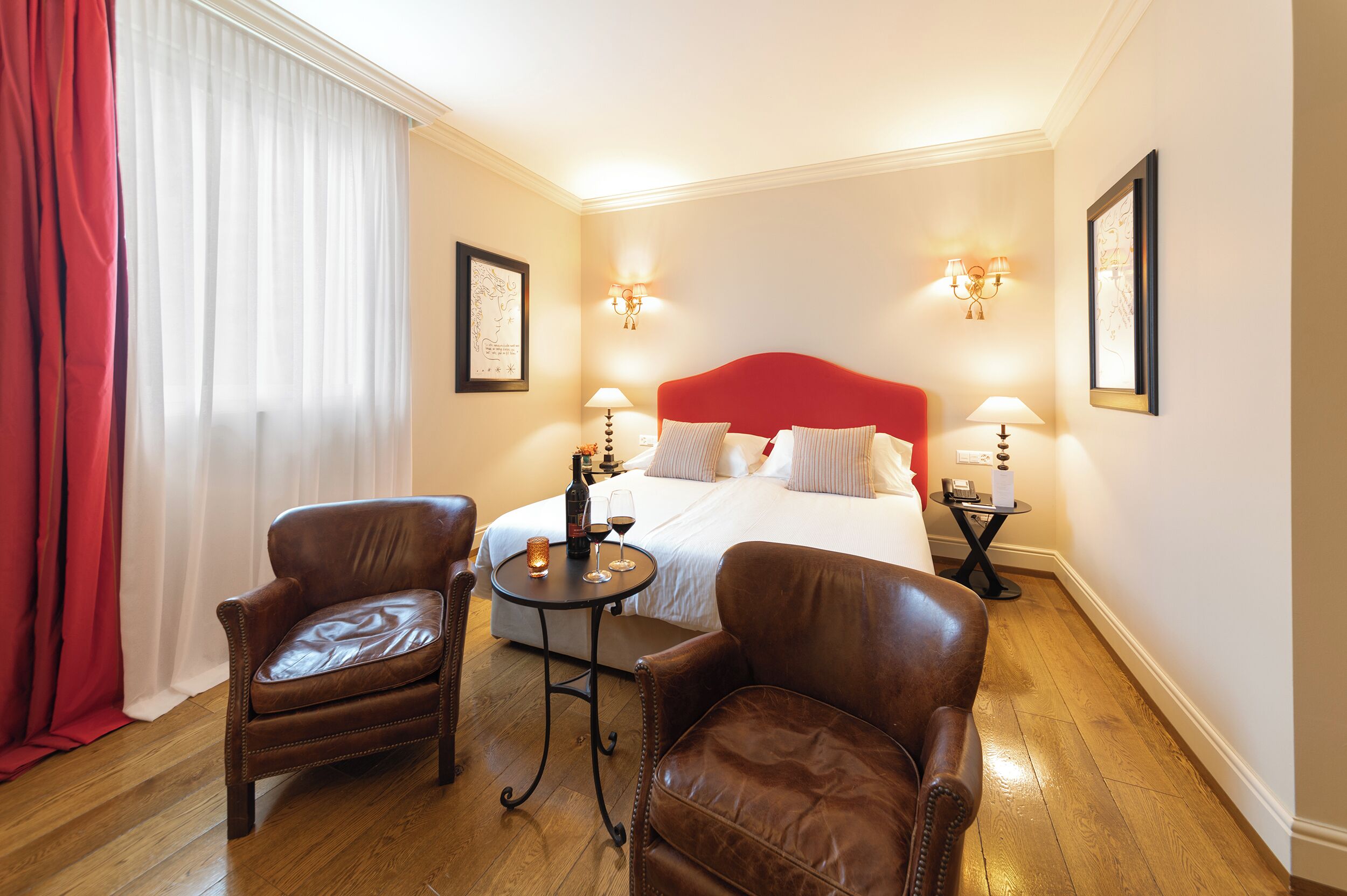 A hotel room with a large bed, two leather chairs, a small table with a bottle and glasses, and two wall-mounted lamps.