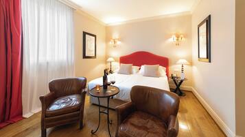 A hotel room with a large bed, two leather chairs, a small table with a bottle and glasses, and two wall-mounted lamps.