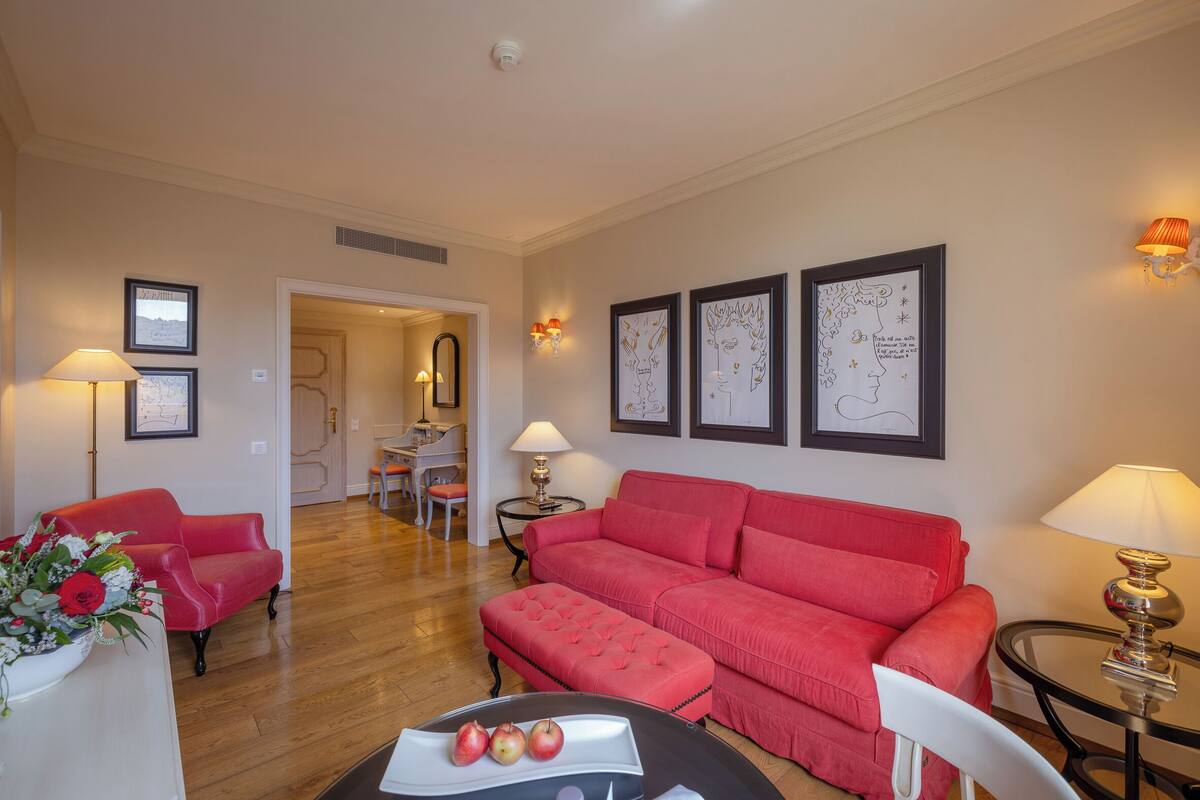 A living room with a red sofa, a white armchair, a small round table with a tray of apples, and a vase of flowers. There are framed pictures on the wall and a lamp on a side table.
