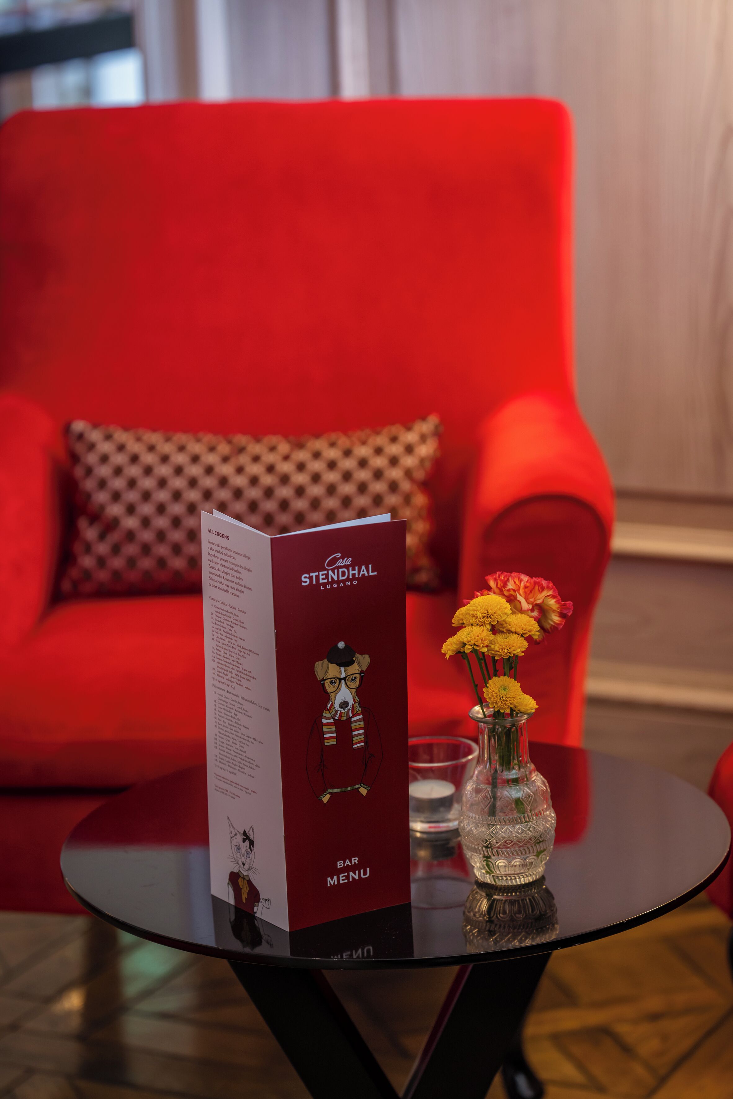 A red sofa with a patterned pillow, a glass table with a menu, and a vase with yellow flowers.