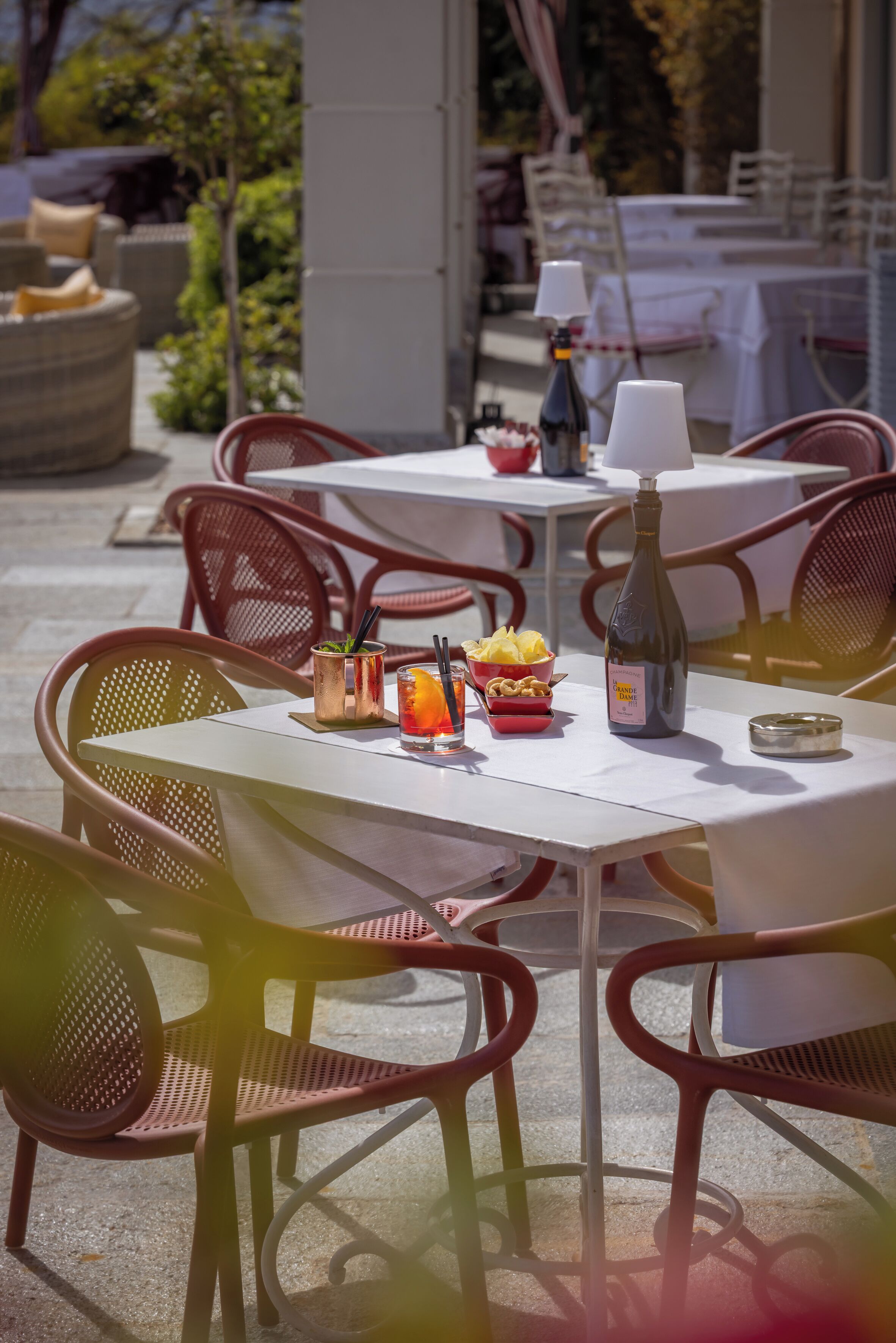 An outdoor dining area with tables and chairs, a bottle of wine, a drink with an orange slice, and a bowl of snacks.