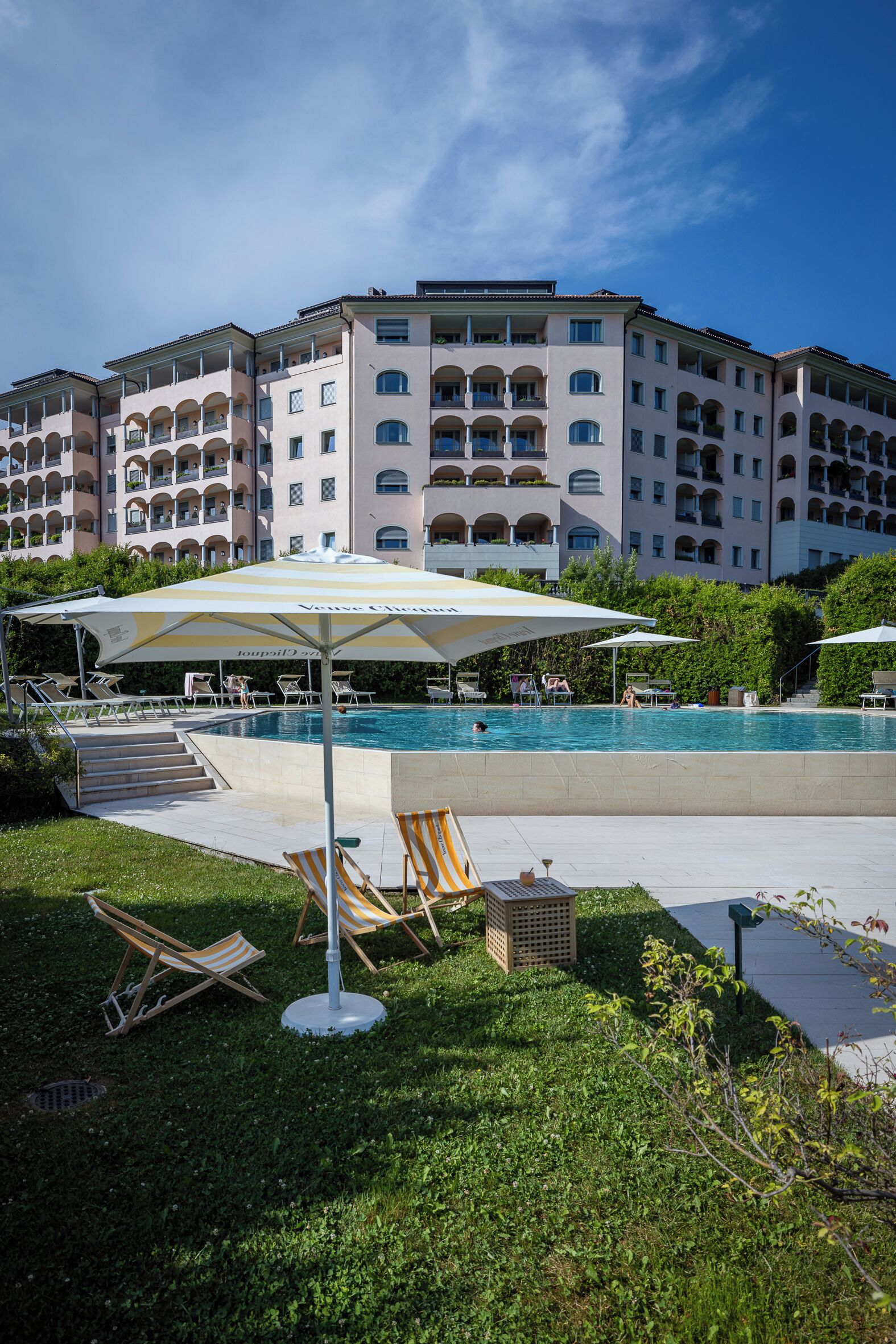 Ein Poolbereich mit Liegestühlen und Sonnenschirmen, umgeben von einem Gebäude mit mehreren Balkonen.