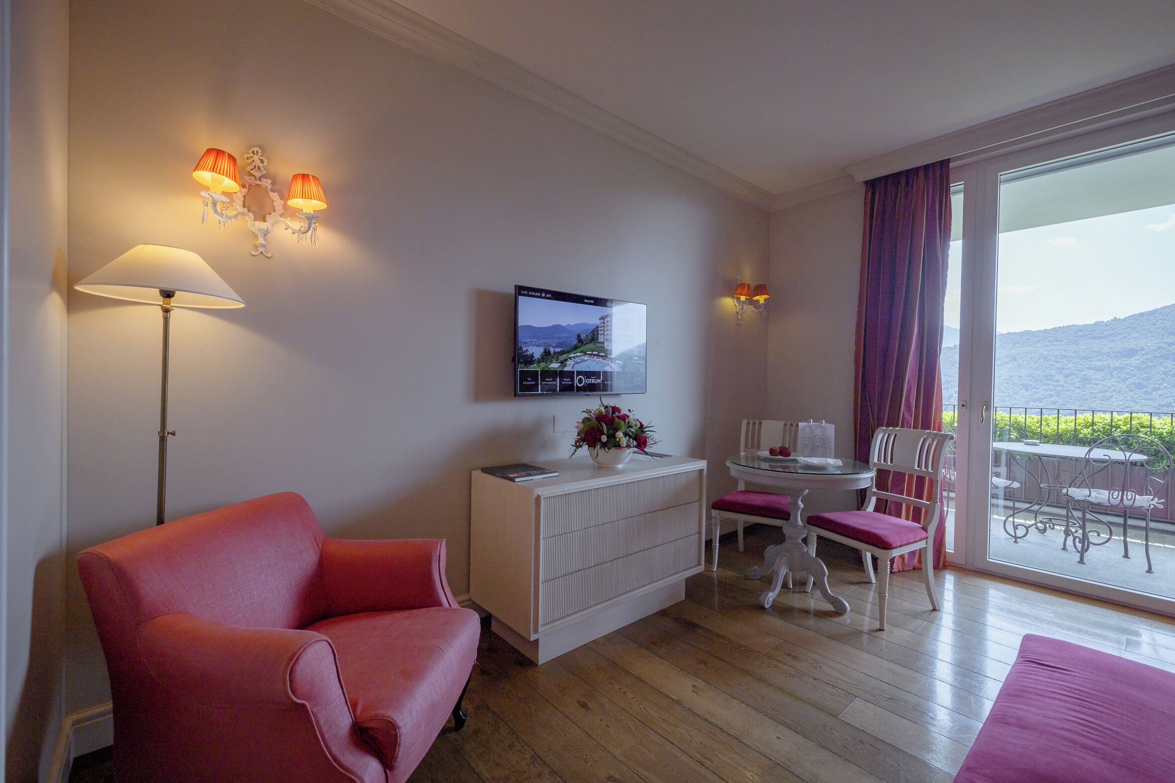 Een kamer met een rode bank, een wit kastje, een televisie, een eettafel met stoelen, en een balkon met uitzicht op zee.