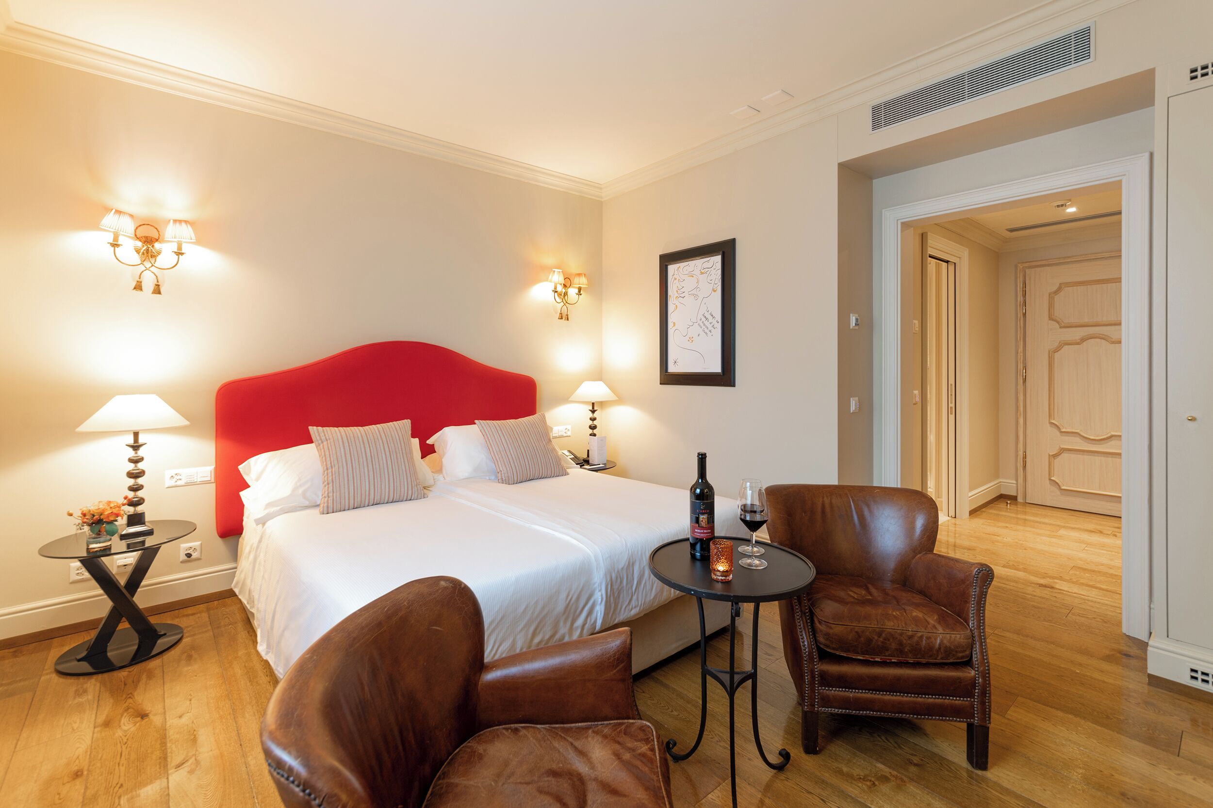 A hotel room with a red headboard bed, a small round table with a bottle and glass, two brown leather chairs, and a nightstand with a lamp and flowers.