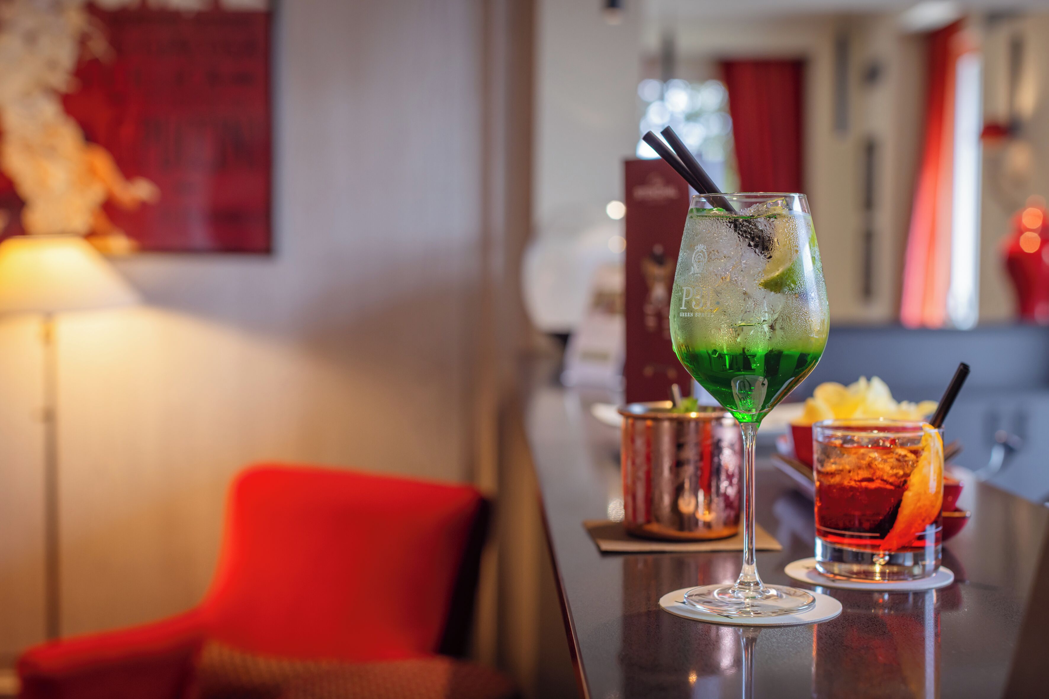 A bar area with a glass of green cocktail, a red cocktail, and a red chair.