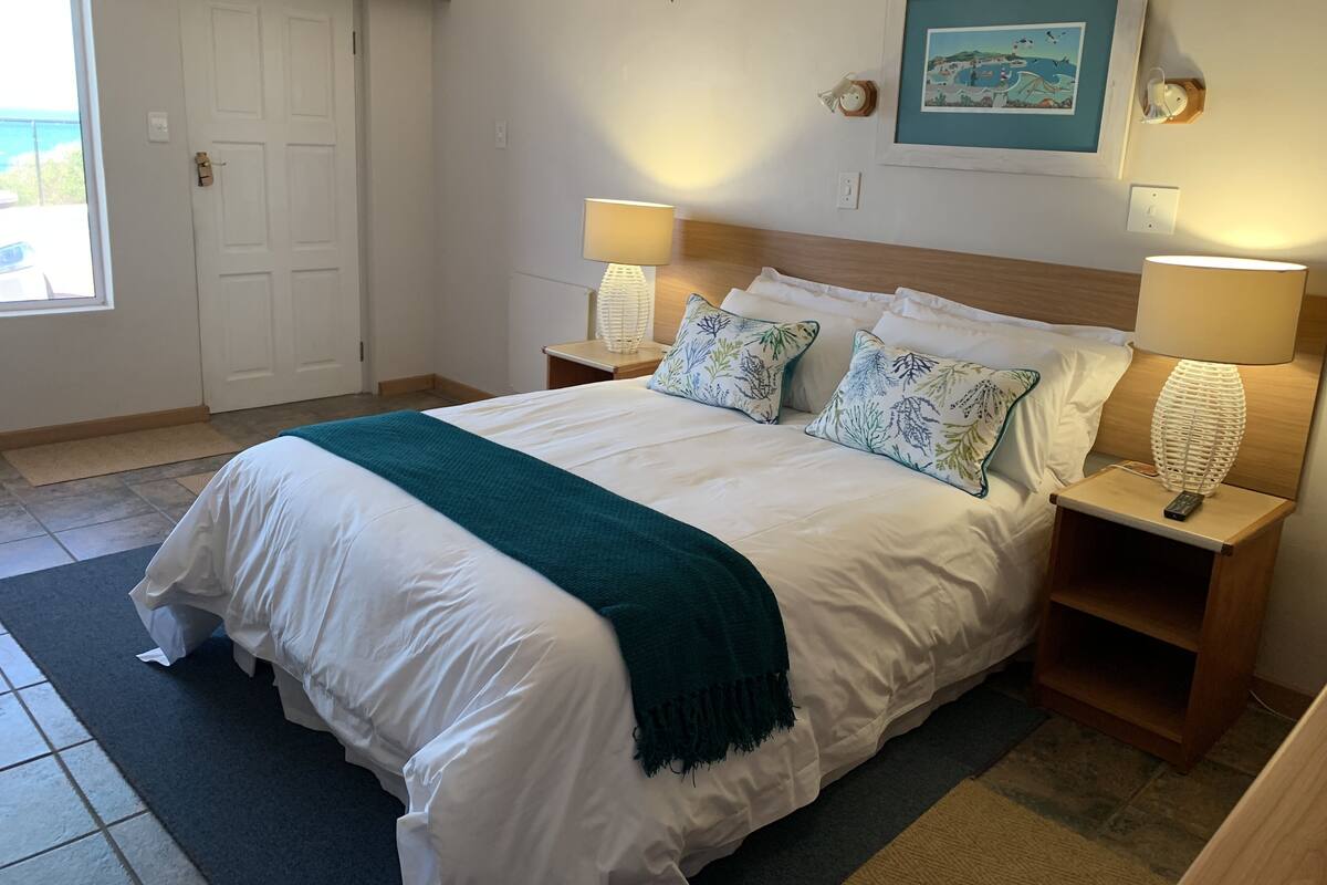 A neatly made bed with white linens and a teal throw, flanked by two bedside tables with lamps, a framed picture on the wall, and a door leading to a balcony.