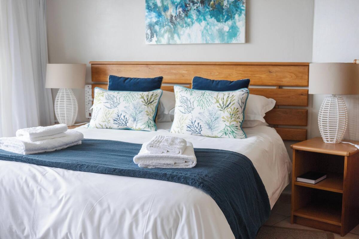 A hotel room with a bed, two pillows, a bedside table, a lamp, and a wall painting.