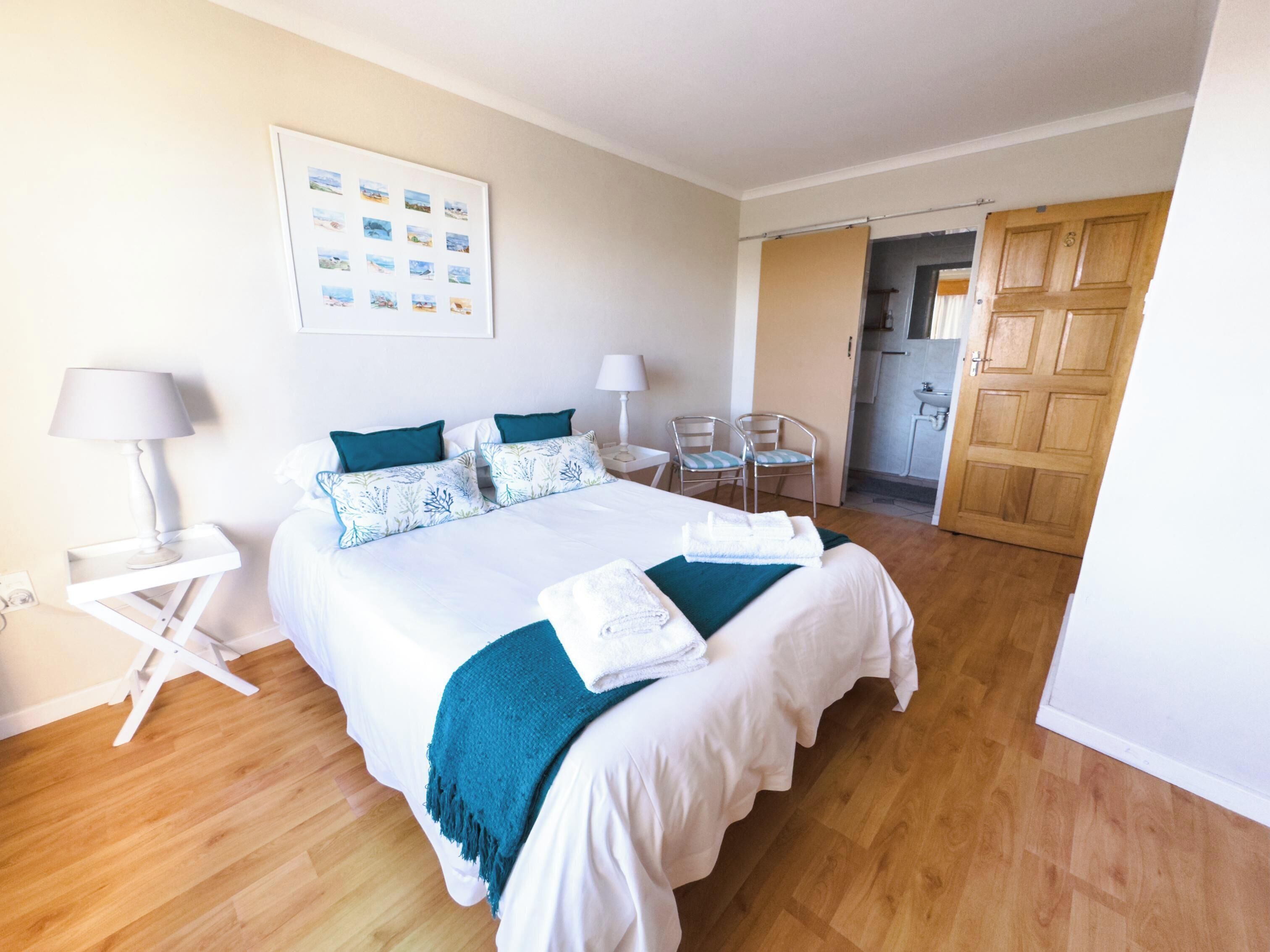 A hotel room with a large bed, two bedside tables, a desk, chairs, a kitchen area, and a door.