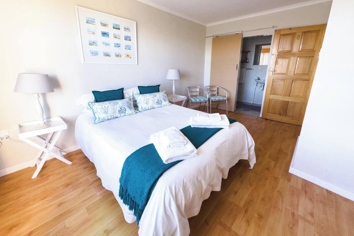 A hotel room with a large bed, two bedside tables, a desk, chairs, a kitchen area, and a door.
