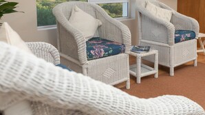 A room with wicker chairs and a small table, a view of greenery outside, and a wooden cabinet. - Aire del Mar Guest House (Kleinbaai)