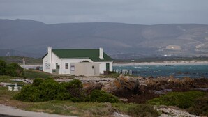 A white building with a green roof, located near a rocky shoreline with a view of the sea and distant hills. - Aire del Mar Guest House (Kleinbaai)
