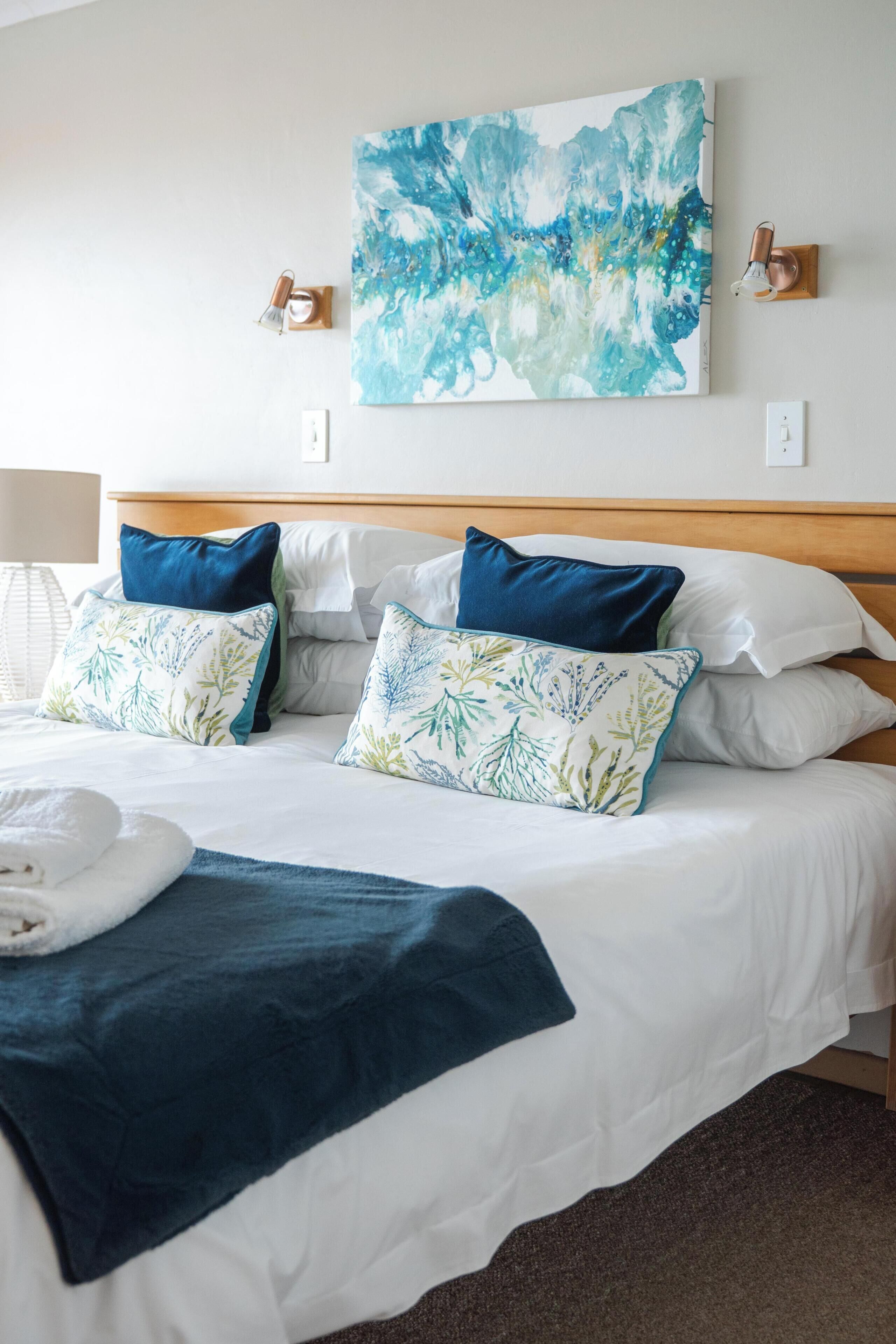 A neatly made bed with white linens and blue and green patterned pillows, a blue throw blanket, and a blue towel on the side.