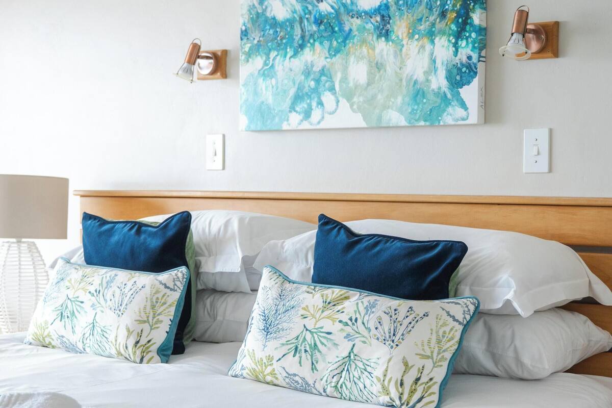 A neatly made bed with white linens and blue and green patterned pillows, a blue throw blanket, and a blue towel on the side.