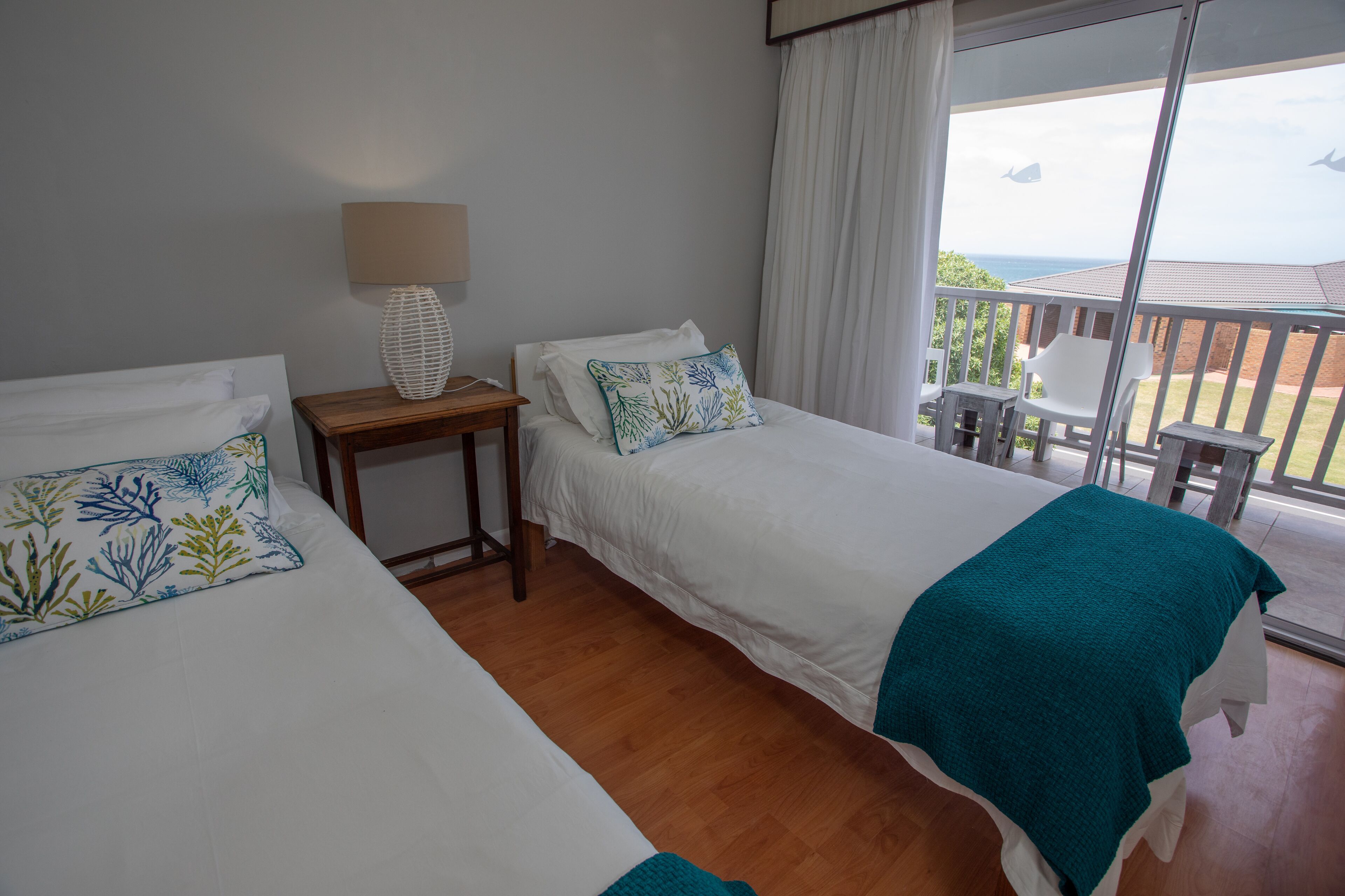 A hotel room with two beds, a small table with a lamp, and a balcony with a view of the sea.