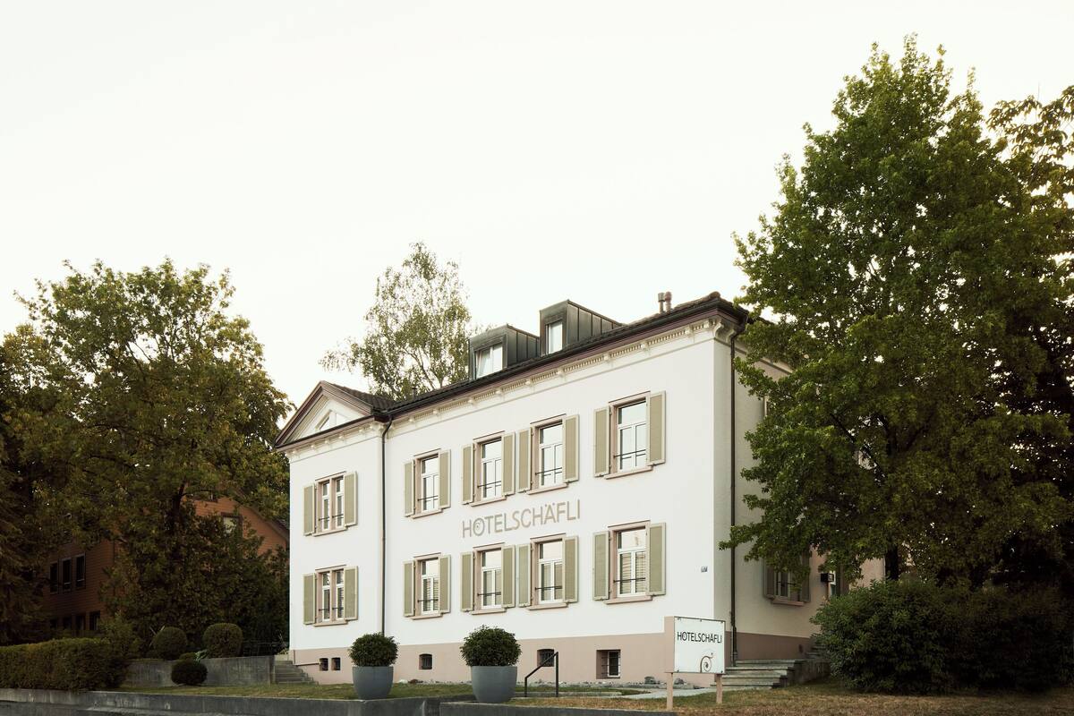 Un edificio de dos pisos con el letrero "HOTELSCHÄFELI" en la fachada, que presenta una disposición simétrica de ventanas y una entrada central.