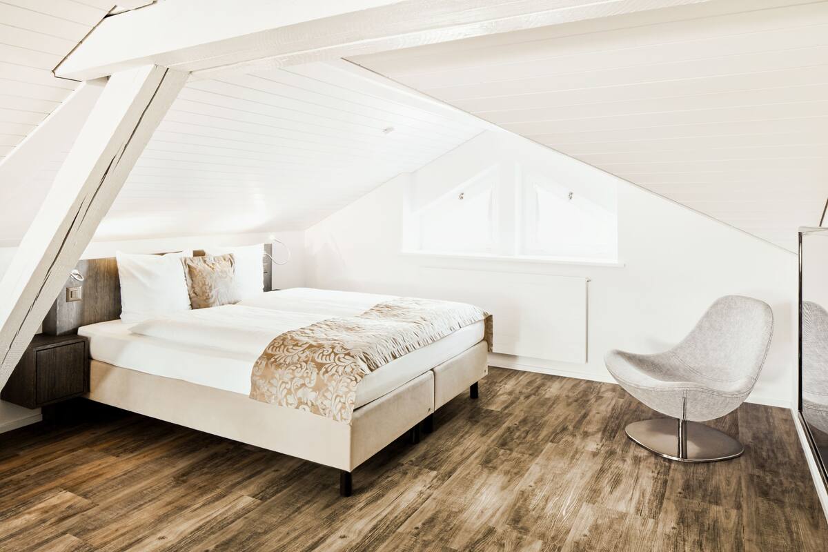 Un dormitorio con techo inclinado, una cama con colcha estampada, una silla moderna y suelo de madera.