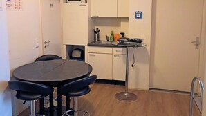 A compact kitchen with a microwave, a sink, and a small countertop. There is a round table with four bar stools and a door with an emergency exit sign. - Pension Puschkin Berlin (Berlin)