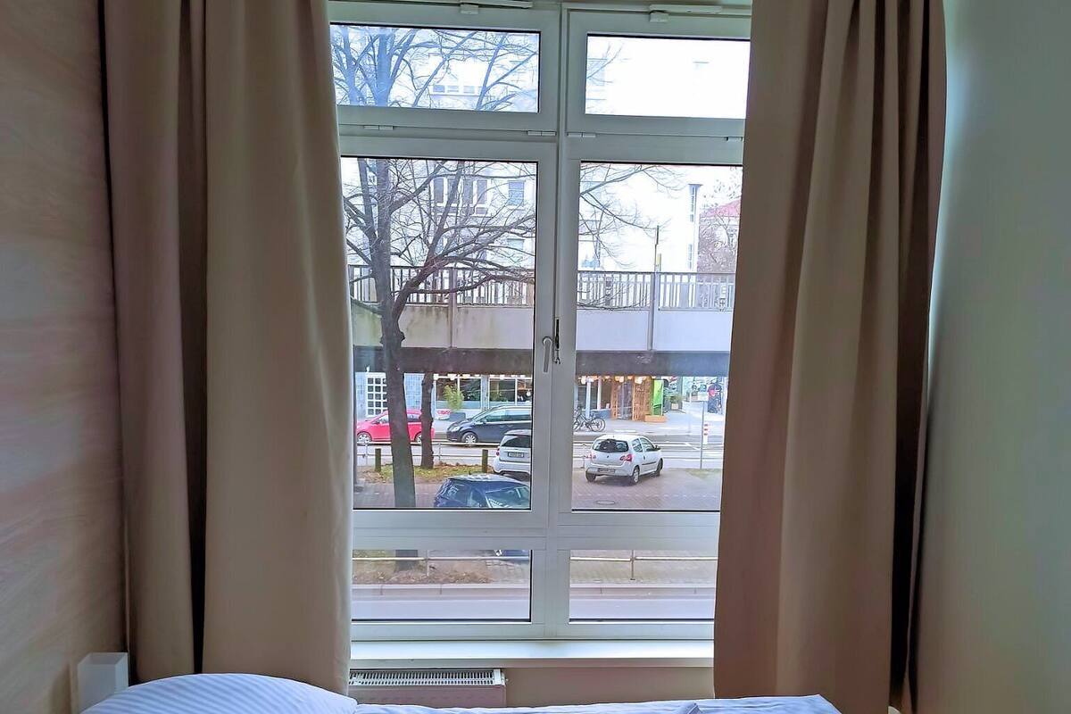 A hotel room with a bed, beige curtains, and a window view of a street with cars and buildings.