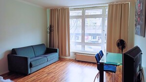 A room with a sofa, a chair, a table, a TV, and a window with curtains. - Pension Puschkin Berlin (Berlin)