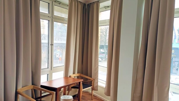 A hotel room with a bed, a wooden table with chairs, a radiator, and large windows with curtains. - Pension Puschkin Berlin (Berlin)