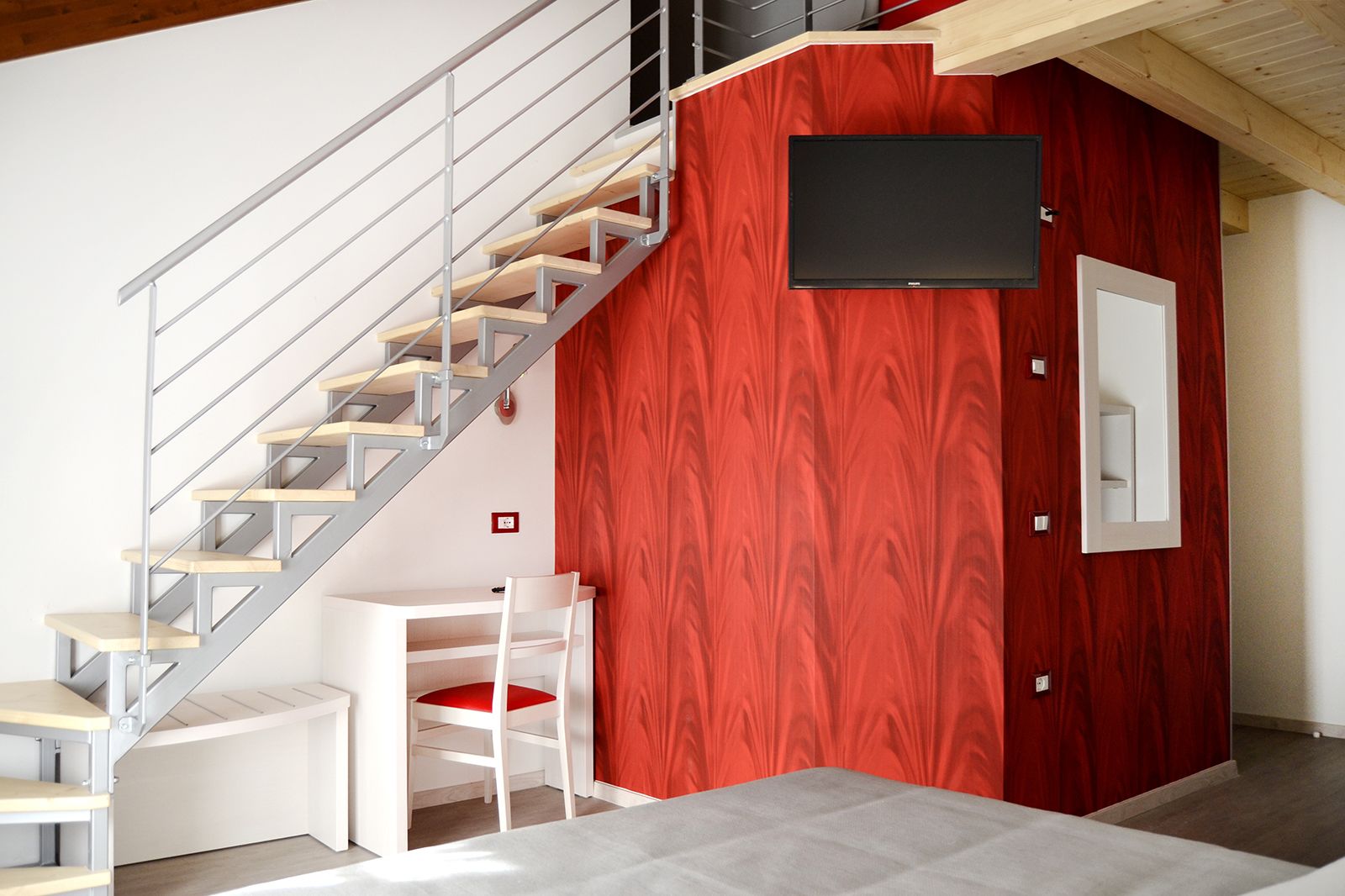 A modern interior with a staircase, a red accent wall, a mounted TV, and a white desk with a chair.
