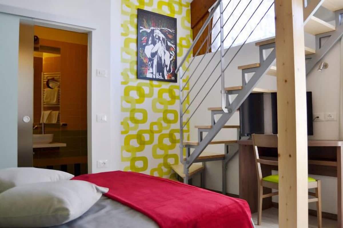 A bedroom with a bed, a red blanket, a wooden chair, a desk, and a staircase.