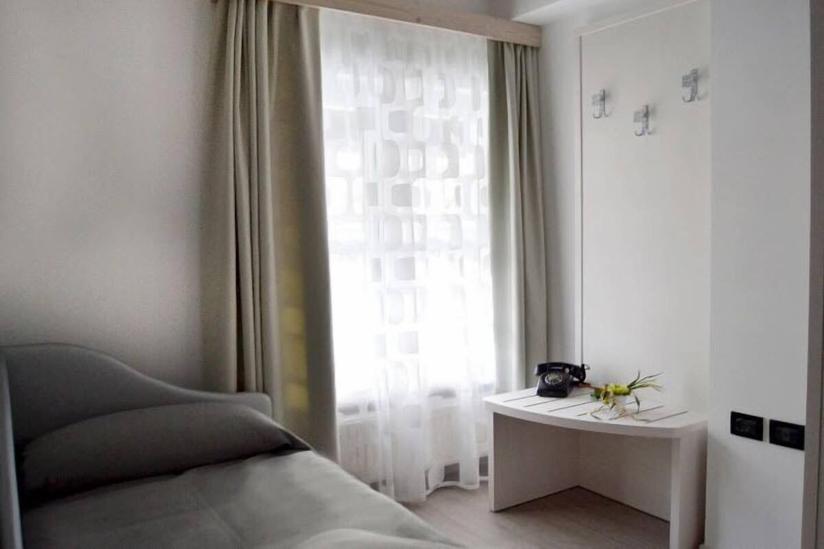 A modern hotel room with a bed, a desk with a telephone and a vase of flowers, and a window with sheer curtains.